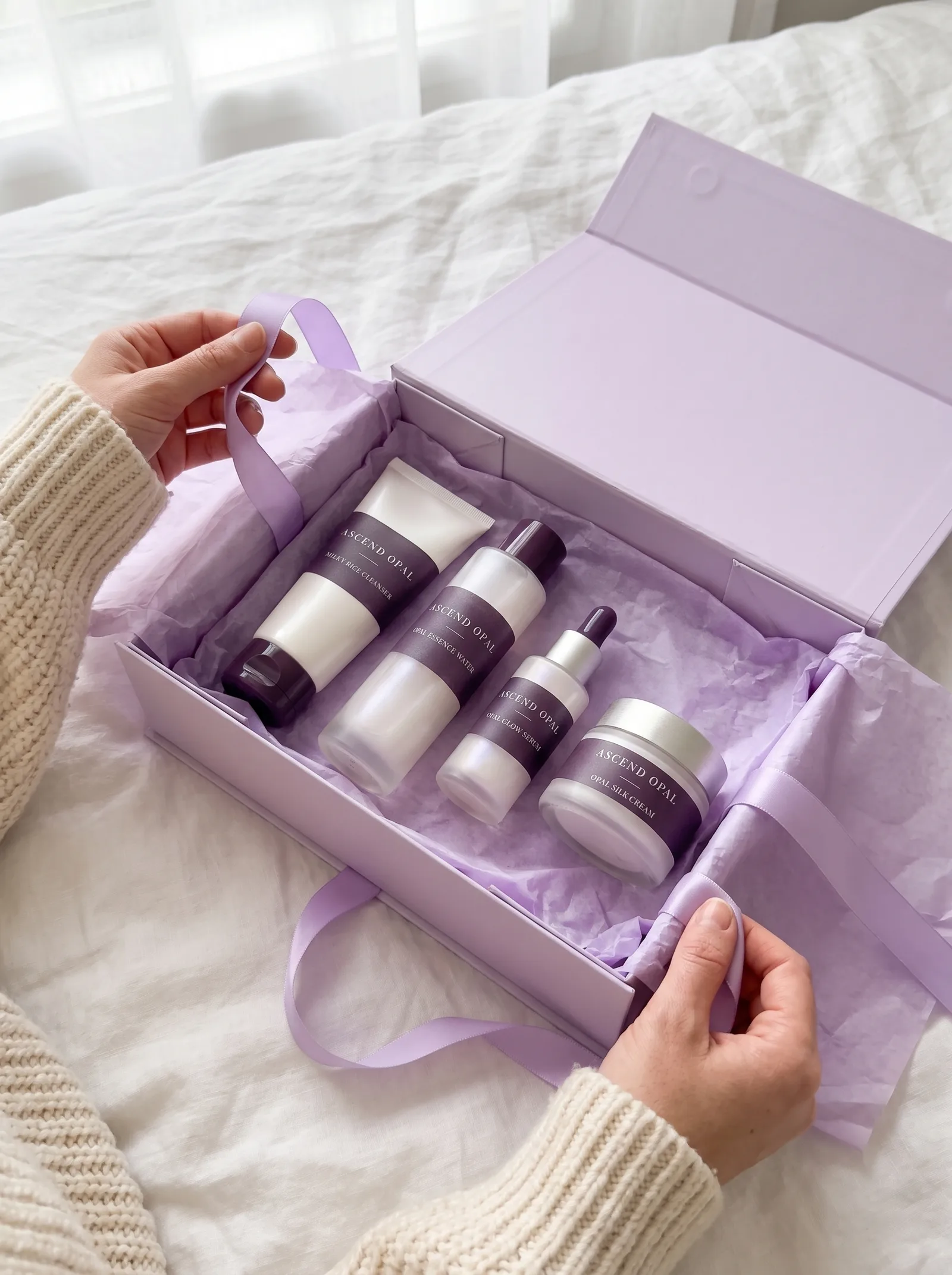 The Opal Set in an open lilac gift box on a linen bed, a hand lifting the ribbon